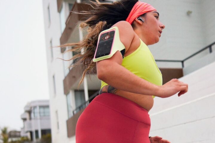 Woman running outside, wearing a phone armband. with Peloton app showing on the screen